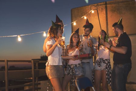 Téléchargez les photos : Groupe de jeunes amis faisant une fête d'anniversaire sur le toit d'un immeuble, chantant une chanson et soufflant une bougie - en image libre de droit