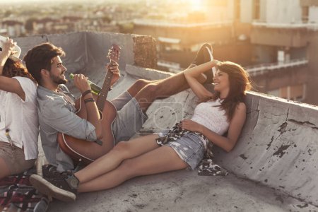 Photo for Young people chilling out and partying on a building rooftop. Focus on the guy playing the guitar - Royalty Free Image
