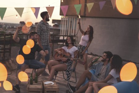 Photo for Young friends having fun at a summertime rooftop party, playing the guitar, singing, dancing and chilling out. Focus on the girls in the middle - Royalty Free Image