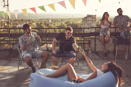 Foto de Grupo de jóvenes amigos divirtiéndose en la fiesta en la azotea, haciendo barbacoa y disfrutando de los calurosos días de verano. Concéntrate en los hombres sentados - Imagen libre de derechos
