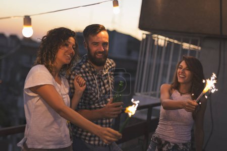Téléchargez les photos : Groupe de jeunes amis s'amusant à une fête sur le toit, chantant, dansant et saluant avec des étincelles. Concentrez-vous sur le gars - en image libre de droit