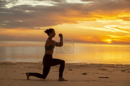 Téléchargez les photos : Jeune femme active faisant de l'exercice sur la plage tôt le matin, faisant des squats fendus et profitant d'un beau lever de soleil sur l'eau - en image libre de droit