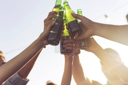Photo for Group of young friends having fun at rooftop party, drinking beer, making a toast and enjoying hot summer days. Selective focus on the beer bottles - Royalty Free Image