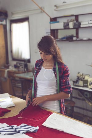 Téléchargez les photos : Jeune couturière coupant le tissu avec des ciseaux, faisant le nouveau modèle de vêtements - en image libre de droit