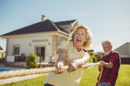 Photo for Senior couple having fun playing tug of war game, spending sunny summer day outdoors; group of elderly friends participating in rope pulling competition at retirement home - Royalty Free Image