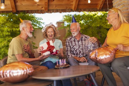 Foto de Grupo de alegres amigos mayores divirtiéndose en una fiesta de cumpleaños, usando sombreros de fiesta y sosteniendo globos mientras el anfitrión abre regalos - Imagen libre de derechos
