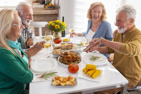 Foto de Amigos mayores celebrando Acción de Gracias juntos en casa durante la cena tradicional, anfitriones cortando el pollo y entreteniendo a los invitados - Imagen libre de derechos