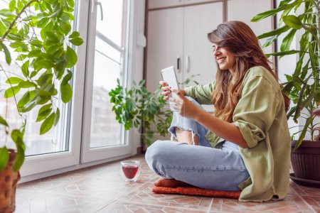 Téléchargez les photos : Femme passe son temps libre à se détendre à la maison, assis à son balcon étage à boire du thé et à lire un ebook à l'aide d'un ordinateur tablette - en image libre de droit