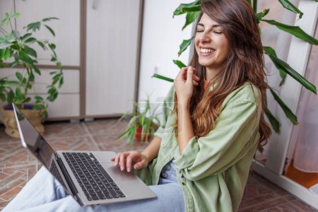 Téléchargez les photos : Belle jeune femme assise sur son balcon tenant ordinateur portable dans ses genoux tout en travaillant à la maison - en image libre de droit