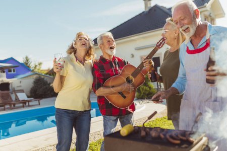 Foto de Grupo de alegres amigos mayores teniendo una fiesta de barbacoa junto a la piscina, reunidos alrededor de la parrilla, tocando la guitarra, cantando, asando carne y divirtiéndose - Imagen libre de derechos