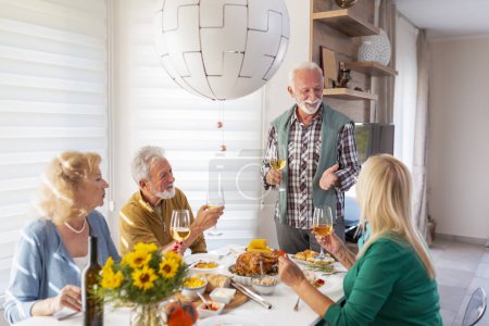 Foto de Dos parejas mayores se reunieron alrededor de la mesa, celebrando Acción de Gracias, cenando juntos en casa y levantando copas de vino, haciendo un brindis, presentando un discurso - Imagen libre de derechos