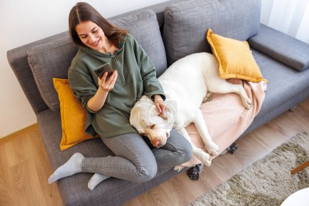 Téléchargez les photos : Joyeux jeune femme se détendre et profiter du temps libre à la maison, dactylographier un message texte à l'aide d'un téléphone intelligent avec son chien de compagnie couché sur ses genoux - en image libre de droit