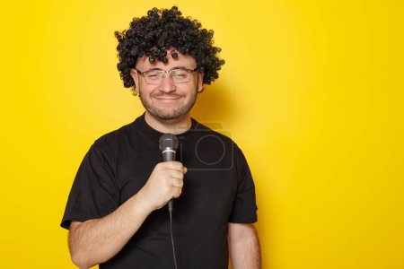 Téléchargez les photos : Portrait d'homme portant perruque, T-shirt noir et lunettes tenant micro et chantant isolé sur fond jaune avec espace de copie - en image libre de droit