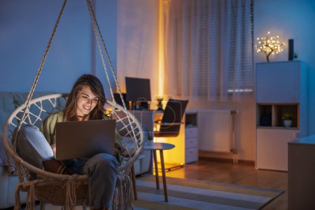 Téléchargez les photos : Femme appréciant le temps libre à la maison, assis dans la chaise suspendue dans le salon en utilisant un ordinateur portable - en image libre de droit