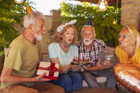 Foto de Grupo de ancianos alegres divirtiéndose en una fiesta de cumpleaños, usando sombreros de fiesta, sosteniendo globos y soplando velas en un pastel de cumpleaños - Imagen libre de derechos