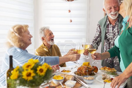 Téléchargez les photos : Deux couples de personnes âgées se sont réunis pour le dîner de Thanksgiving, hôte exprimant sa gratitude à sa femme pour avoir préparé un bon repas et porter un toast en levant des verres de vin, accueillant les invités - en image libre de droit