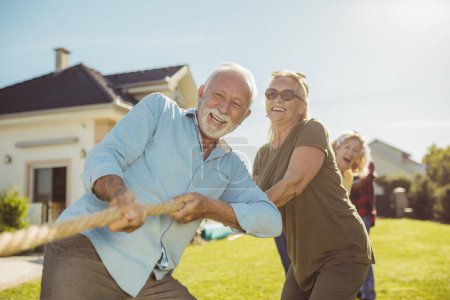 Photo for Senior people having fun playing tug of war, spending sunny summer day outdoors; group of elderly friends having fun participating in rope pulling competition at retirement home - Royalty Free Image