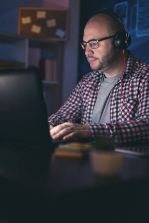Foto de Hombre con auriculares trabajando en el ordenador portátil en la oficina en casa tarde en la noche; desarrollador web trabajando horas extras - Imagen libre de derechos