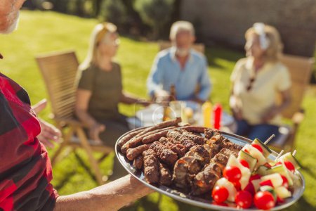 Photo for Group of cheerful senior friends having an outdoor lunch in the backyard, gathered around the table, host bringing food on a tray, offering it to guests - Royalty Free Image