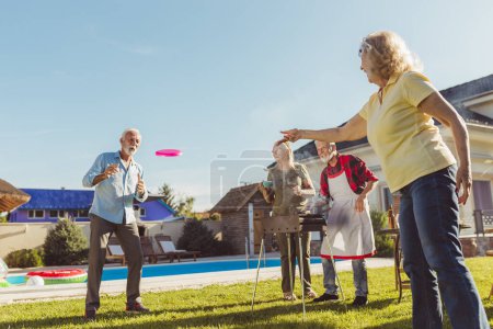 Foto de Grupo de alegres personas mayores activas que tienen una fiesta de barbacoa en el patio trasero, asar carne y verduras y divertirse jugando a tirar y coger - Imagen libre de derechos