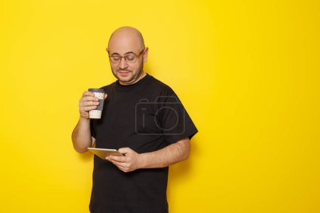Téléchargez les photos : Homme lecture ebook à l'aide d'un ordinateur tablette et boire emporter café isolé sur fond de couleur jaune avec espace de copie - en image libre de droit