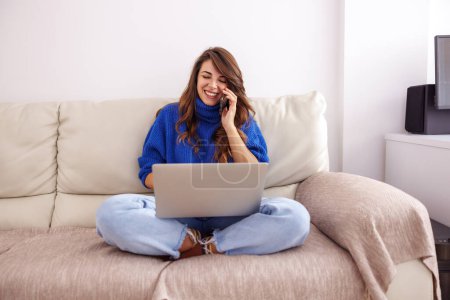 Photo for Young woman sitting on couch, having phone conversation and using laptop computer while working remotely from home - Royalty Free Image