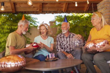Téléchargez les photos : Groupe de personnes âgées gaies s'amusant à une fête d'anniversaire, portant des chapeaux de fête, tenant des ballons et ouvrant des cadeaux - en image libre de droit