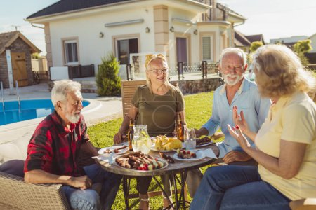 Photo for Group of elderly people eating lunch outdoors by the swimming pool, gathered around the table, eating, drinking and relaxing on a sunny summer day - Royalty Free Image
