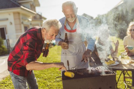 Photo for Elderly neighbors having fun while grilling meat in the backyard, relaxing outdoors on sunny spring day - Royalty Free Image