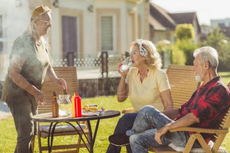 Photo for Group of cheerful elderly people having an outdoor lunch in the backyard, gathered around the table, drinking beer and lemonade and relaxing on a sunny summer day - Royalty Free Image
