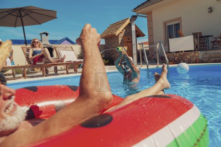 Foto de Grupo de personas mayores que se divierten pasando un día caluroso de verano en la piscina, tomando el sol, flotando en el colchón de aire y salpicándose el agua entre sí, enfriándose y relajándose. - Imagen libre de derechos