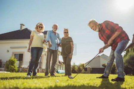 Foto de Grupo de amigos mayores que se divierten jugando mini golf en el césped del patio trasero, pasando un día soleado de verano al aire libre - Imagen libre de derechos