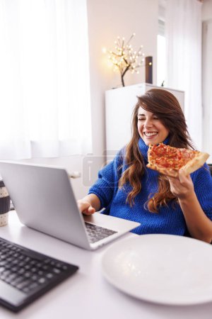 Téléchargez les photos : Développeur web femme assis à son bureau à la maison, prendre une pause tout en travaillant à distance de la maison boire du café et manger de la pizza - en image libre de droit