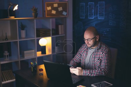 Photo for Man sitting at his desk in home office, typing text message using smart phone while working late at night - Royalty Free Image