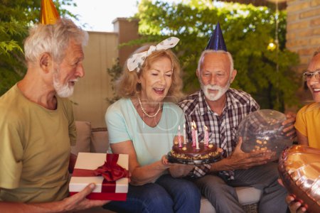 Photo for Group of cheerful elderly people having fun at a birthday party, wearing party hats, holding balloons and blowing out candles at birthday cake - Royalty Free Image