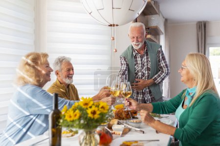 Foto de Dos parejas mayores se reunieron alrededor de la mesa, celebrando Acción de Gracias, cenando juntos en casa y levantando copas de vino, haciendo un brindis, anfitrión dando la bienvenida a los invitados - Imagen libre de derechos