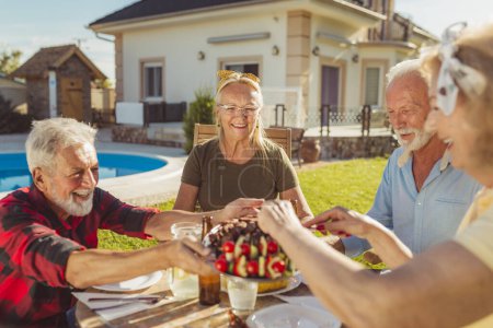 Photo for Group of cheerful senior friends eating lunch in the backyard by the pool, gathered around the table, host serving food on a tray, offering guests - Royalty Free Image