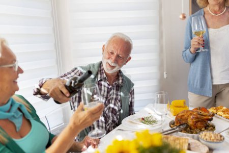 Foto de Grupo de amigos mayores divirtiéndose mientras celebran Acción de Gracias juntos en casa durante la cena tradicional, sirviendo vino y haciendo un brindis - Imagen libre de derechos