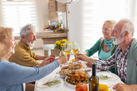 Foto de Dos parejas mayores divirtiéndose mientras celebran Acción de Gracias juntas en casa durante la cena tradicional, levantando copas de vino, haciendo un brindis - Imagen libre de derechos