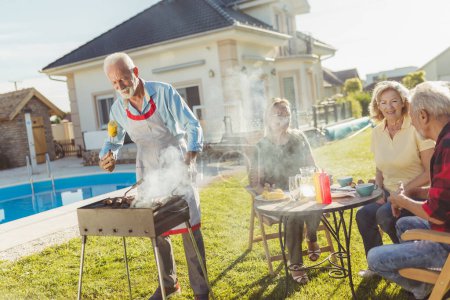 Téléchargez les photos : Des voisins âgés s'amusent à passer une journée ensoleillée en plein air, à faire une fête barbecue dans la cour près de la piscine - en image libre de droit