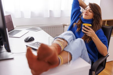 Téléchargez les photos : Programmeuse assise à son bureau à la maison, prenant une pause café, souriant et relaxant tout en travaillant à distance de la maison - en image libre de droit