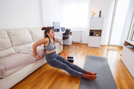 Photo for Active young woman in sportswear doing triceps dips beside the couch while working out at home - Royalty Free Image
