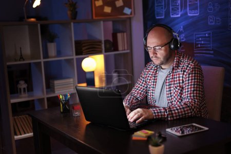 Téléchargez les photos : Homme portant casque travaillant sur ordinateur portable dans le bureau à la maison tard dans la nuit ; développeur web travaillant des heures supplémentaires - en image libre de droit