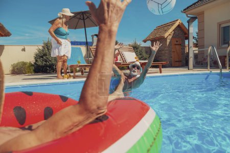 Foto de Dos parejas mayores disfrutando de sus vacaciones de verano juntas, mujeres tomando cócteles y tomando el sol en tumbonas junto a la piscina mientras los hombres juegan voleibol en la piscina - Imagen libre de derechos