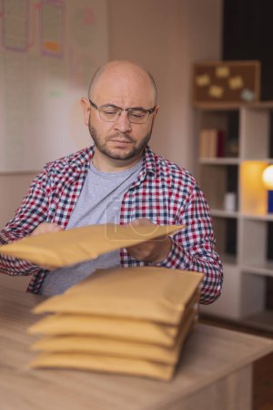 Photo for Small business owner sitting at his desk in home office, packing product for delivery; entrepreneur packaging online store orders for shipping - Royalty Free Image