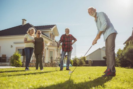 Foto de Grupo de vecinos mayores se reunieron jugando mini golf en el césped del patio trasero, divirtiéndose pasando un día soleado de verano al aire libre - Imagen libre de derechos