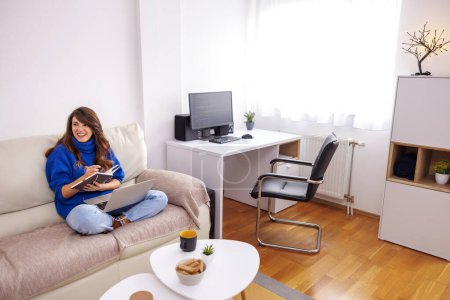 Foto de Mujer sentada en el sofá de la sala de estar sosteniendo el ordenador portátil en su regazo y escribiendo en planificador mientras trabaja desde casa - Imagen libre de derechos