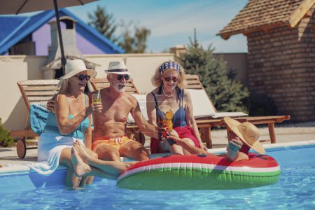 Téléchargez les photos : Groupe de personnes âgées gaies se détendre et bronzer au bord de la piscine pendant les vacances d'été, faire un toast, boire des cocktails et de la bière et s'amuser - en image libre de droit