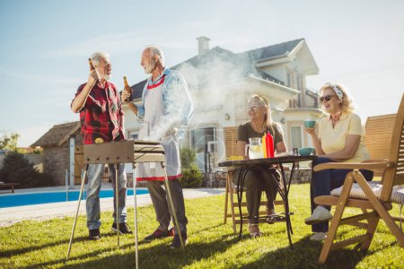 Foto de Amigos mayores divirtiéndose en la fiesta de barbacoa en el patio trasero, hombres asando carne y bebiendo cerveza mientras las mujeres están sentadas y relajándose en el fondo - Imagen libre de derechos