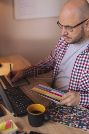 Téléchargez les photos : Designer assis à son bureau à la maison, tenant une palette de couleurs et dessin à l'aide d'un tampon de dessin - en image libre de droit